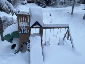 Una estructura de juego de madera cubierta de nieve con un tobogán y columpios, rodeada de árboles nevados y un vehículo al fondo.
