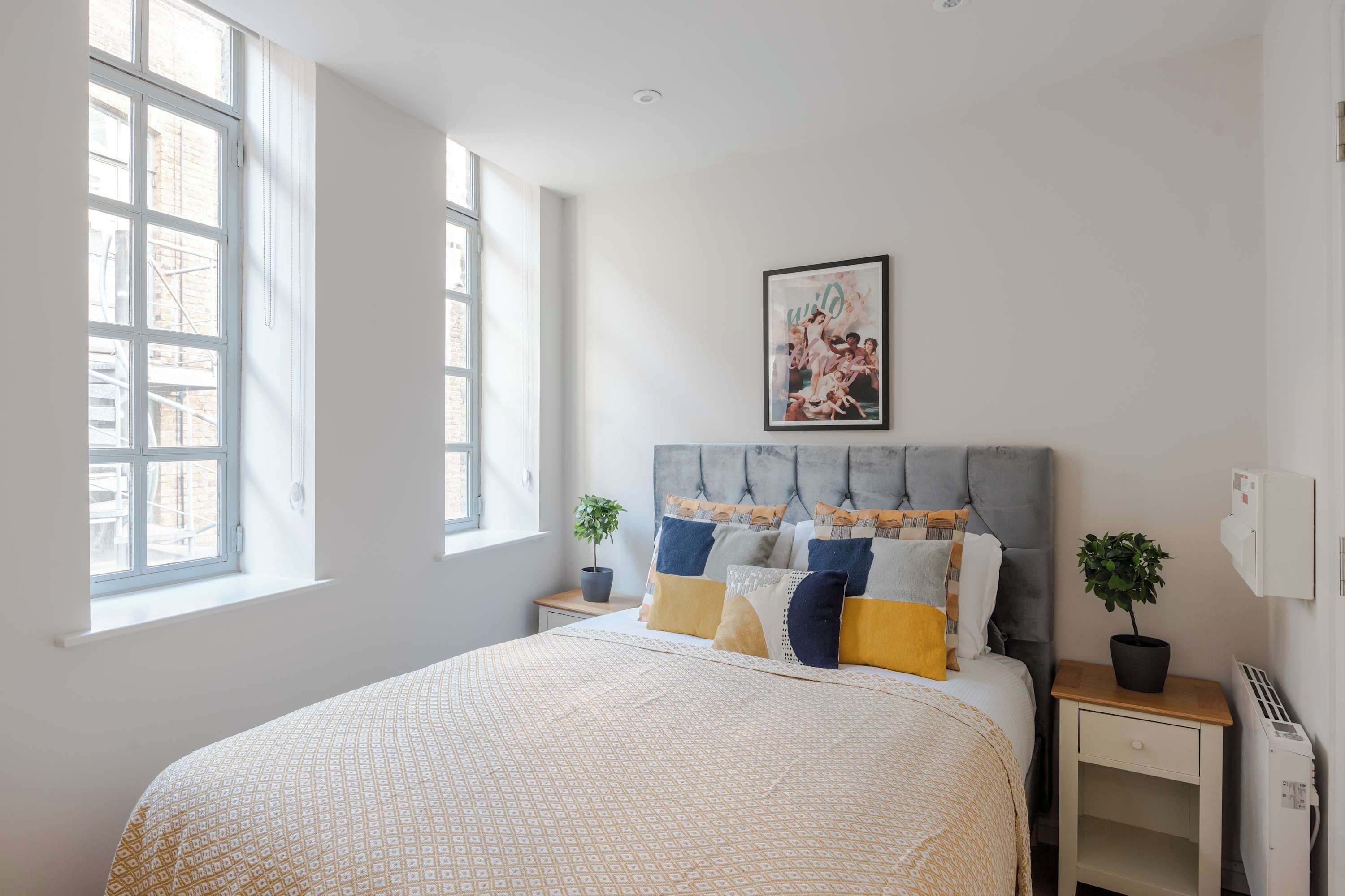 Un dormitorio con una cama, una mesita de noche, una planta en maceta, un cuadro en la pared y un ventanal.
