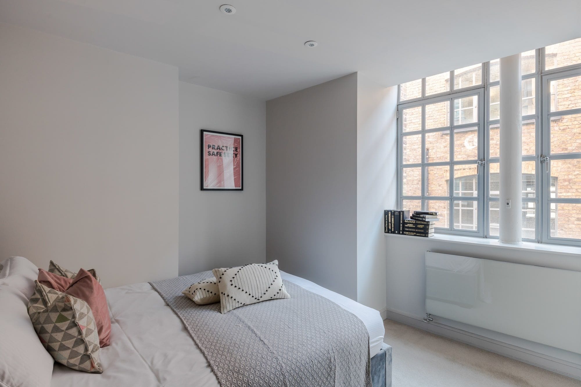 A bedroom with a bed, pillows, a framed poster, a window, and a radiator.
