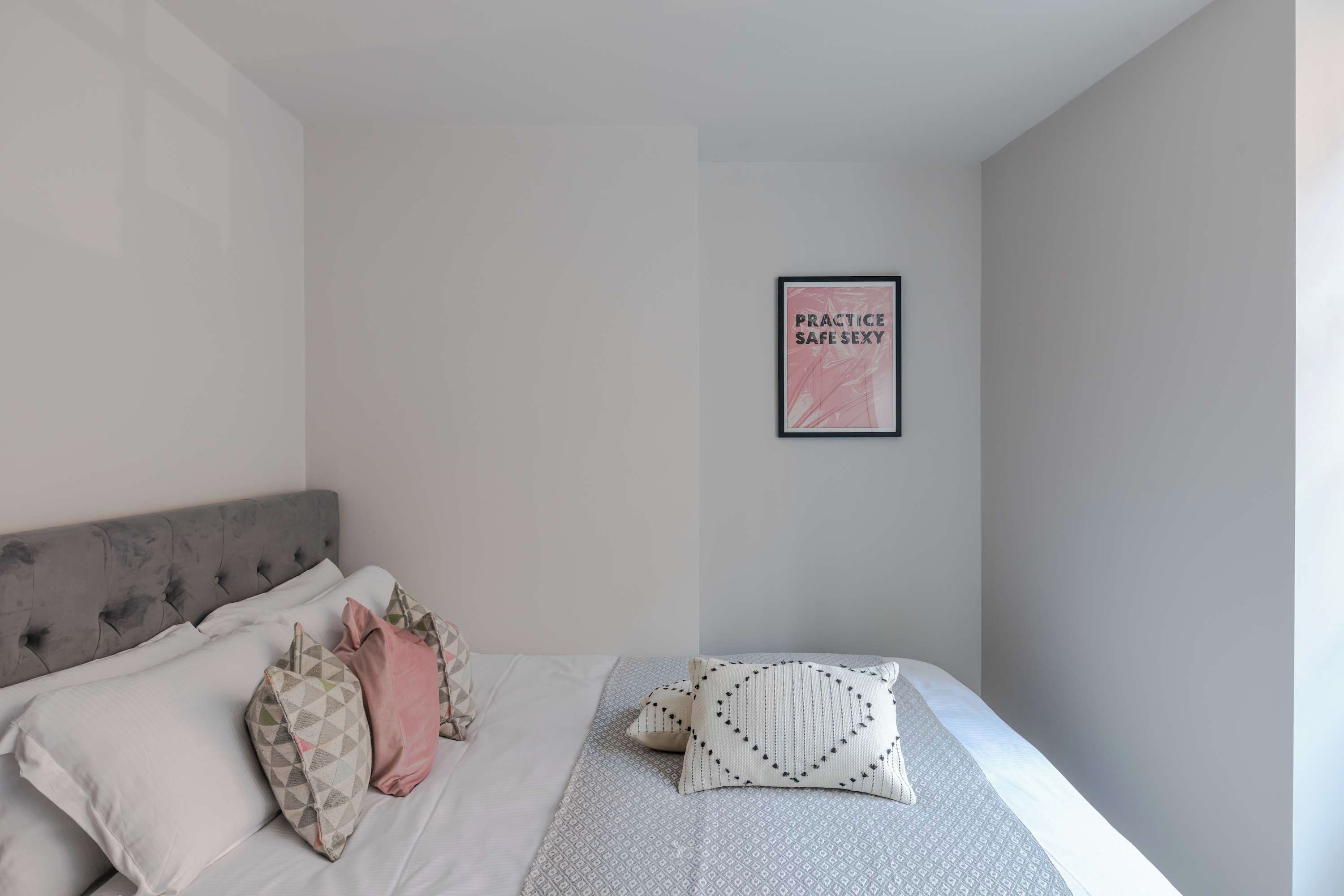 Un dormitorio con un cabecero tapizado gris, una cama con ropa de cama estampada y un cartel enmarcado en la pared.