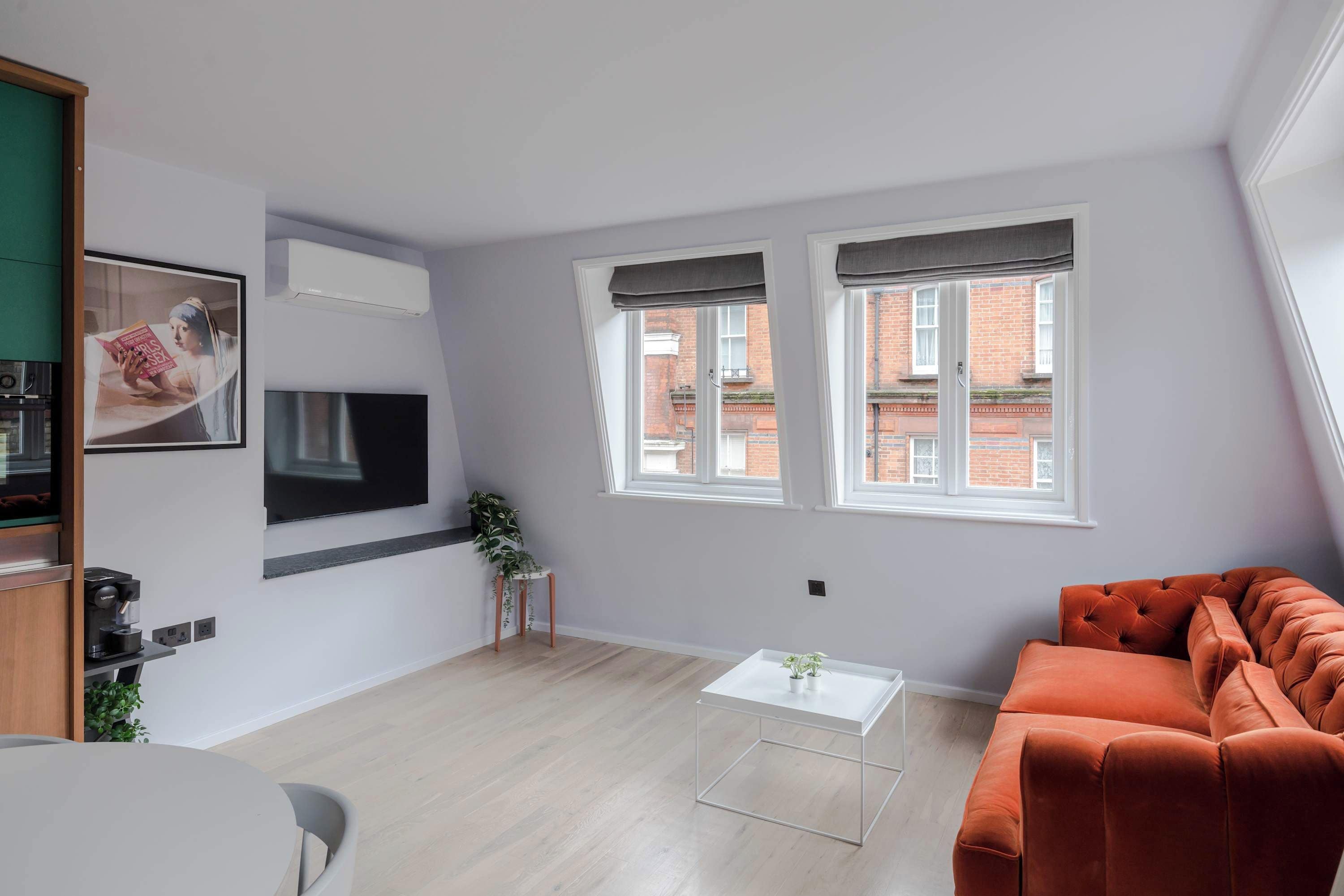 A modern living room with a sofa, a coffee table, and a TV mounted on the wall. There are two windows with curtains, a small plant, and a view of buildings outside.