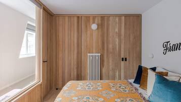 Een slaapkamer met een houten lambrisering, een radiator, een bed met beddengoed met een patroon en een raam met jaloezieën.