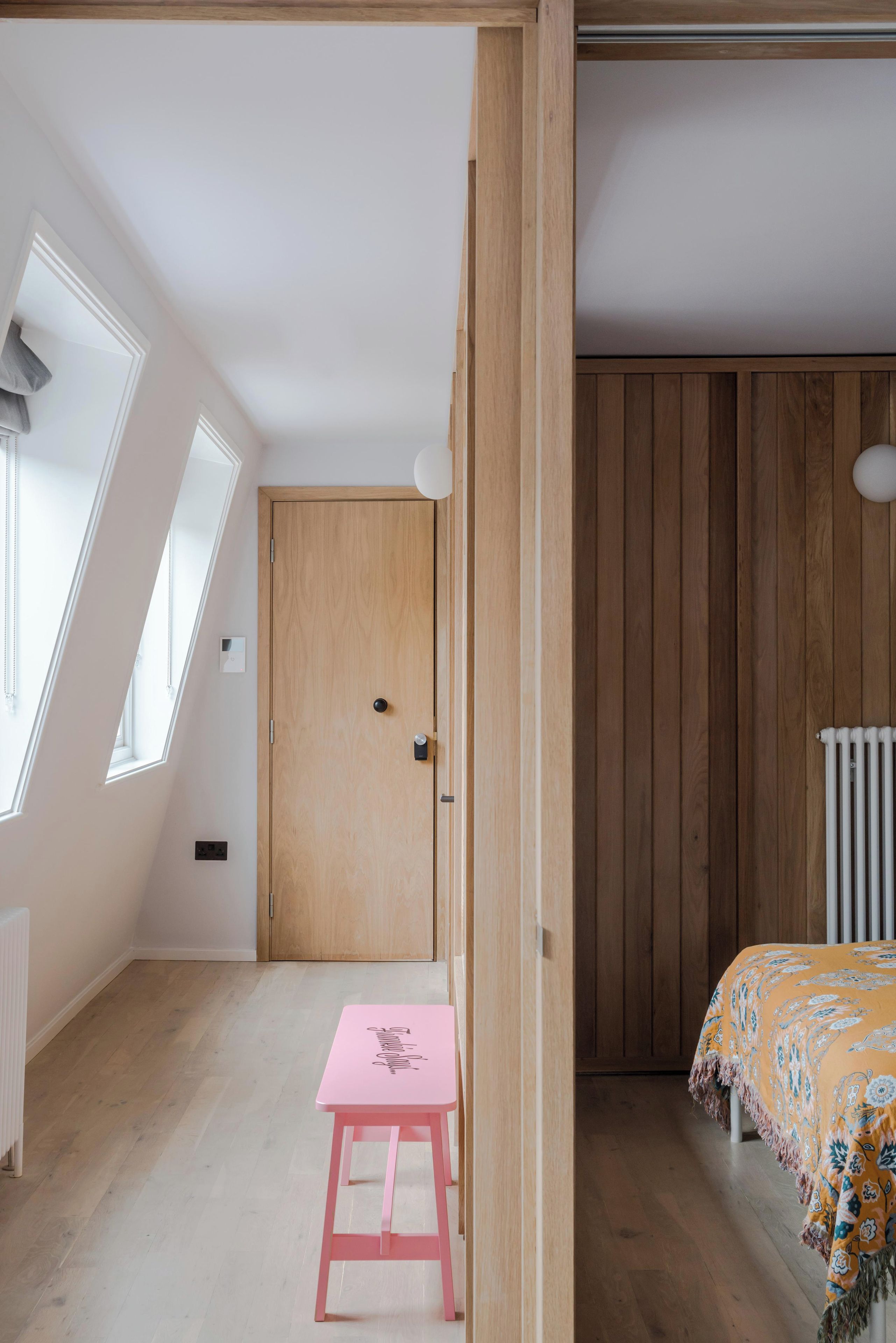 Een kamer met een schuine plafond, een houten deur, een radiator, een bed met een beddegoed met patroon en een roze kruk.