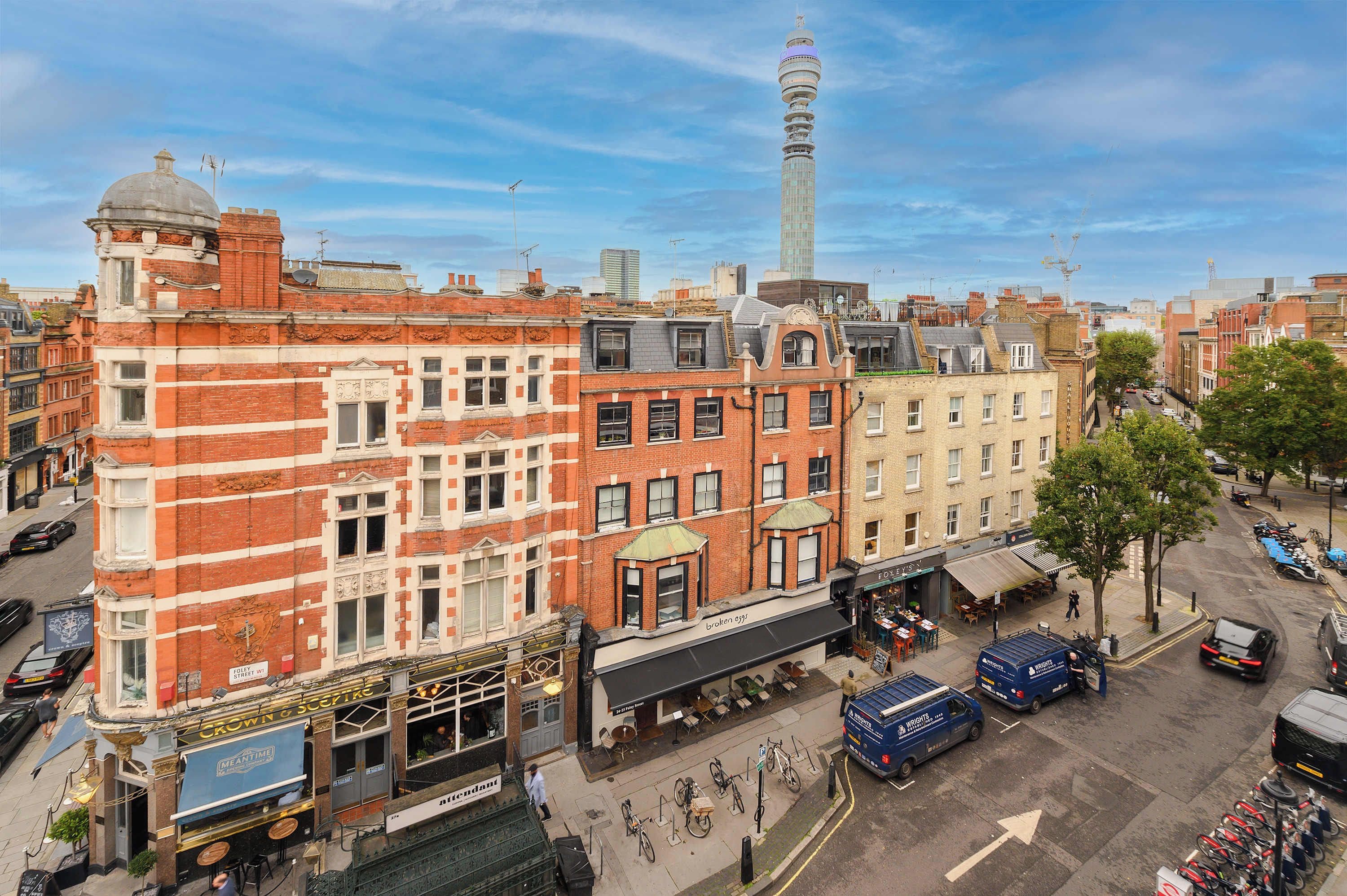 A cityscape with a prominent tower, red brick buildings, and a street with parked cars and bicycles.
