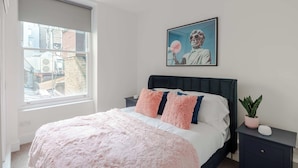 A bedroom with a bed, a nightstand, a potted plant, and a window with a view of buildings. - The Foley Residence - Fitzrovia - by Frankie (London)
