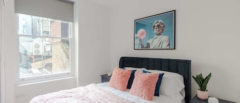 A bedroom with a bed, a nightstand, a potted plant, and a window with a view of buildings.