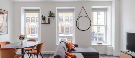 Un salón moderno con un sofá gris, sillones naranjas, una lámpara colgante redonda y grandes ventanales con vistas a la ciudad.