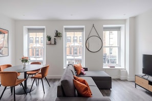 Ein modernes Wohnzimmer mit einem grauen Sofa, orangenen Sesseln, einer runden Pendelleuchte und großen Fenstern mit Blick auf die Stadt.