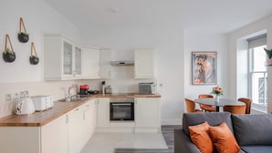 A modern kitchen with white cabinets, a microwave, and a toaster. There is a dining area with a table and chairs, and a gray sofa with orange cushions. - The Foley Residence - Fitzrovia - by Frankie (London)
