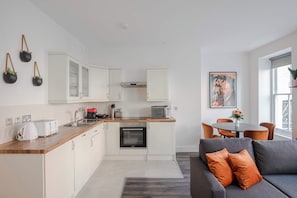 Eine moderne Küche mit weißen Schränken, einer Mikrowelle und einem Toaster. Es gibt eine Essecke mit Tisch und Stühlen sowie ein graues Sofa mit orangefarbenen Kissen.