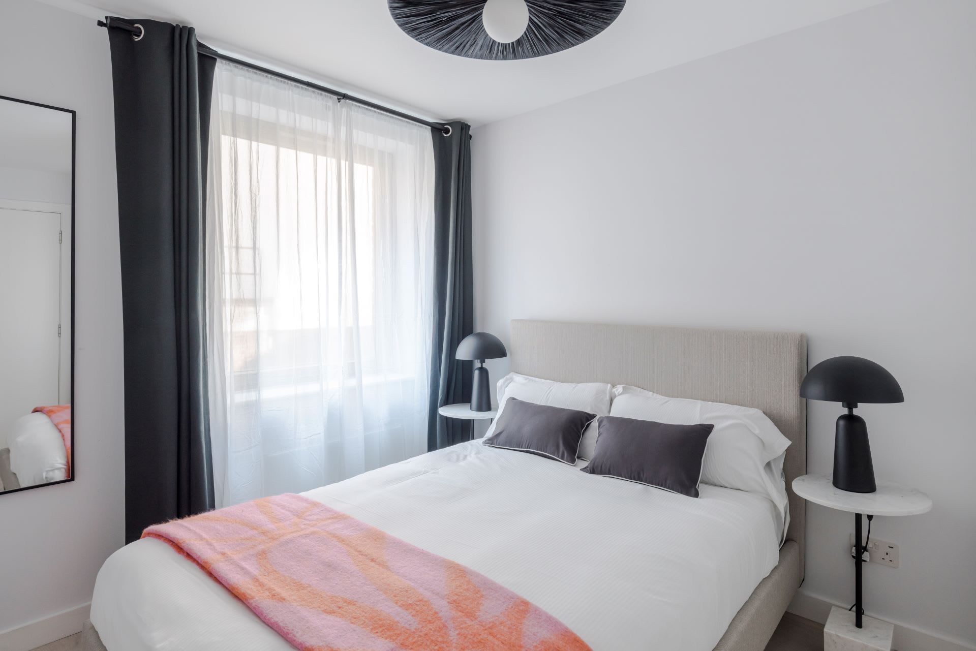A modern hotel room with a large bed, two bedside tables with lamps, a window with sheer curtains, and a ceiling light fixture.