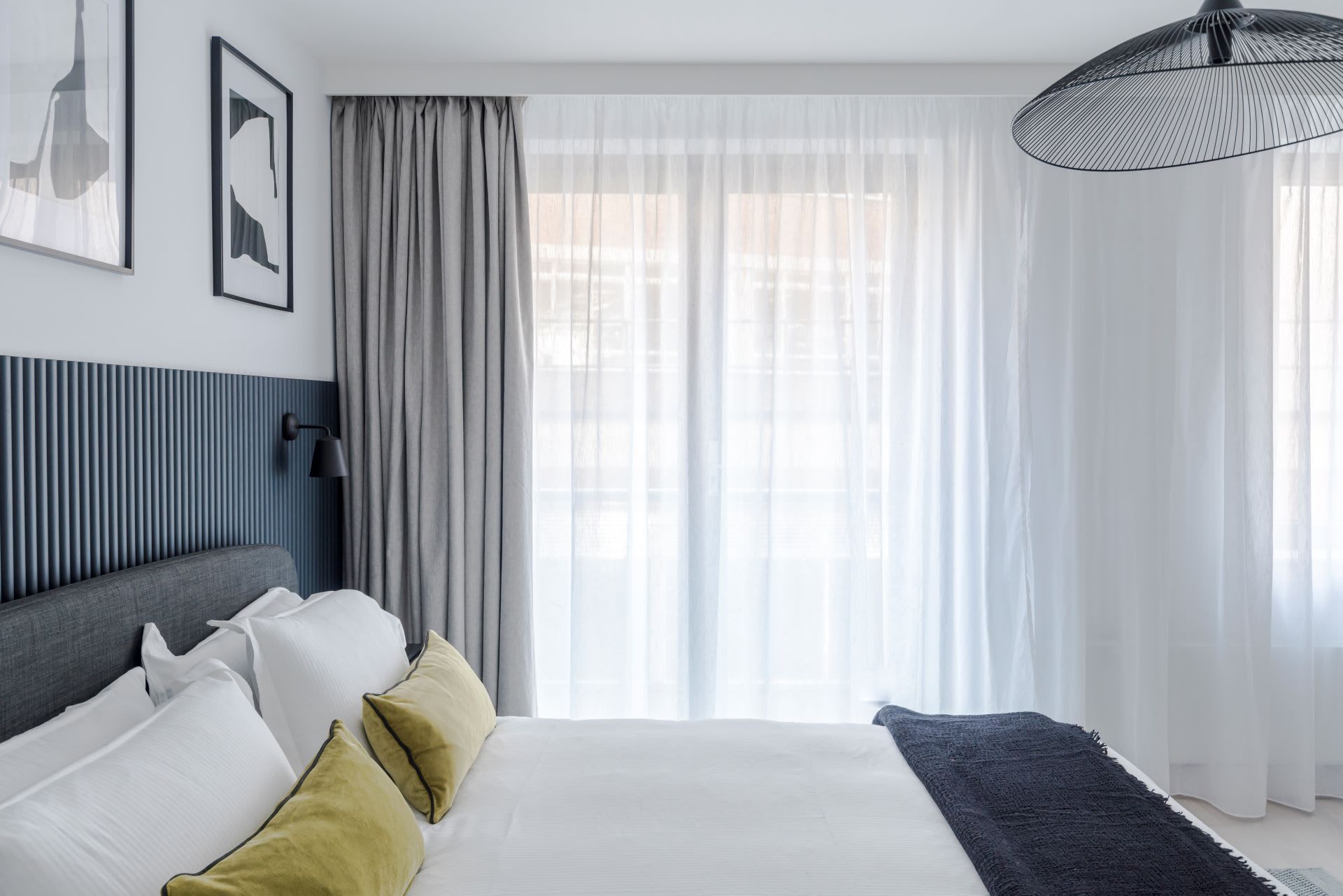 Een moderne hotelkamer met een groot bed, grijze gordijnen, een zwart hanglamp en twee ingelijste kunstwerken aan de muur.