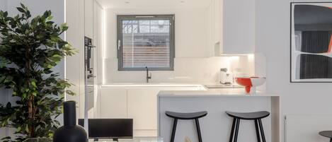 A modern kitchen with white cabinetry, a window with blinds, a white countertop, a red bowl, and two black bar stools.