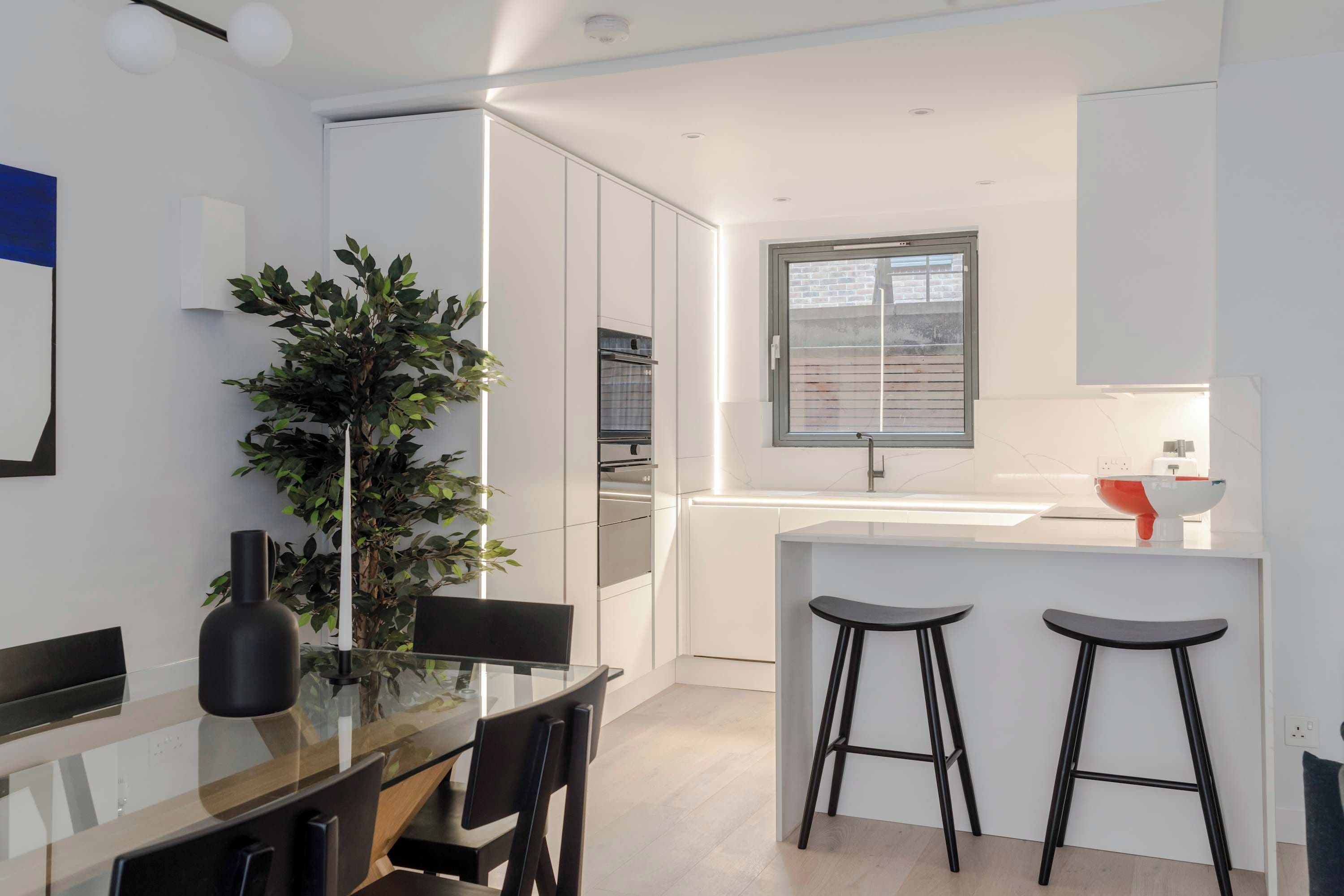 Una cocina moderna con armarios blancos, una isla central con taburetes, una planta en maceta y una ventana con vistas a la ciudad.