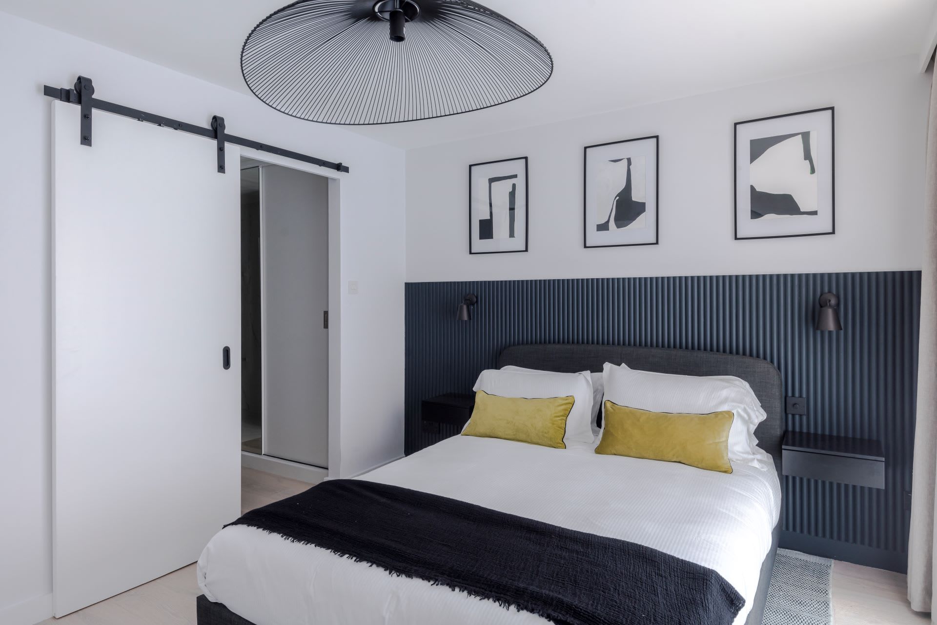 A modern bedroom with a bed, a bedside table, a wall-mounted lamp, and three framed artworks.