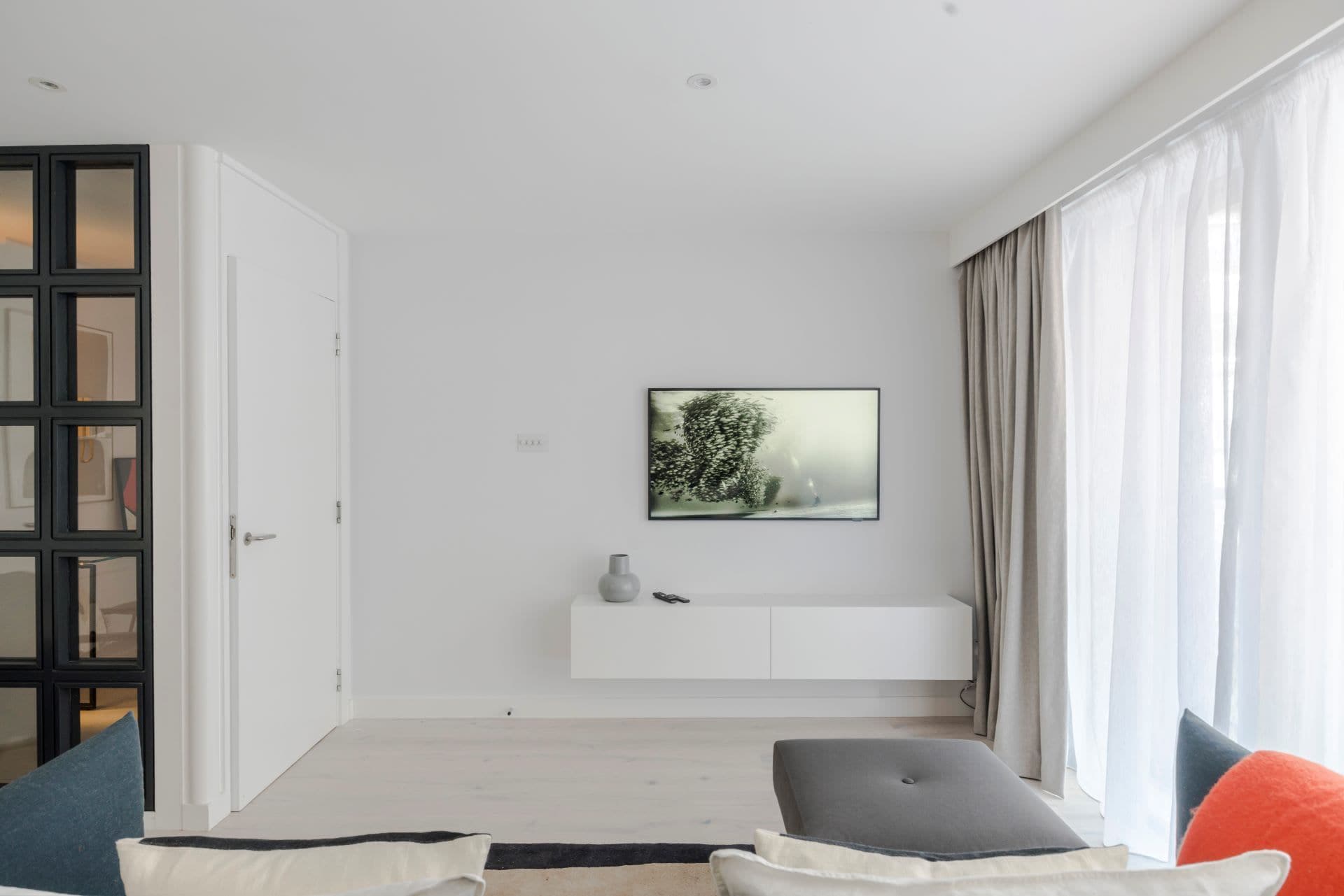 Ein modernes Wohnzimmer mit minimalistischem Design, das eine weiße Wand, ein Wandbild, einen weißen Konsolentisch und ein großes Fenster mit Vorhängen zeigt.