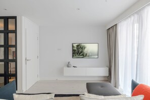 Ett modernt vardagsrum med en minimalistisk design, med en vit vägg, ett upphängt konstverk, ett vitt konsolbord och ett stort fönster med gardiner.