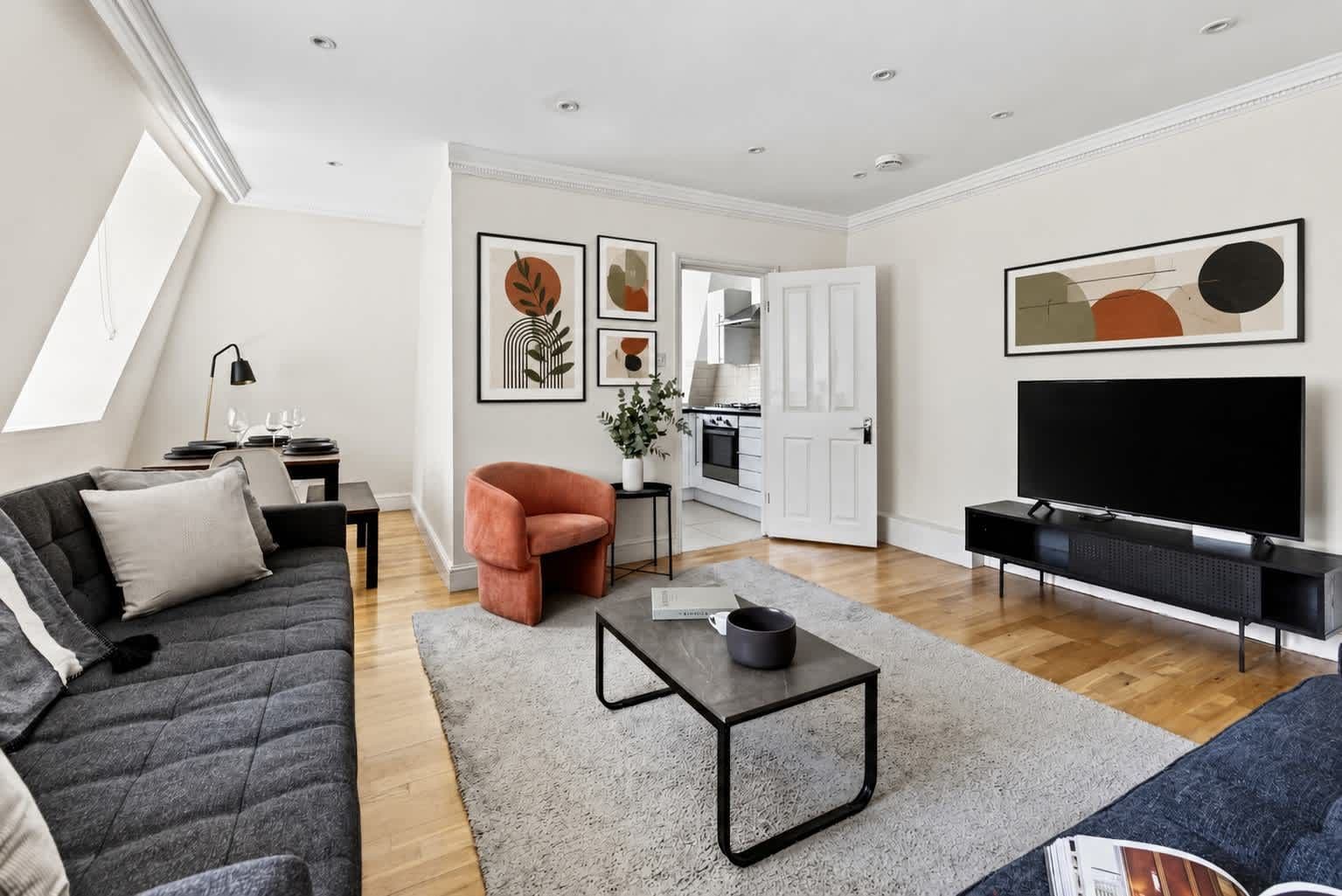 Un salon moderne avec un canapé, une table basse et un téléviseur. On aperçoit un coin cuisine à travers une porte ouverte.