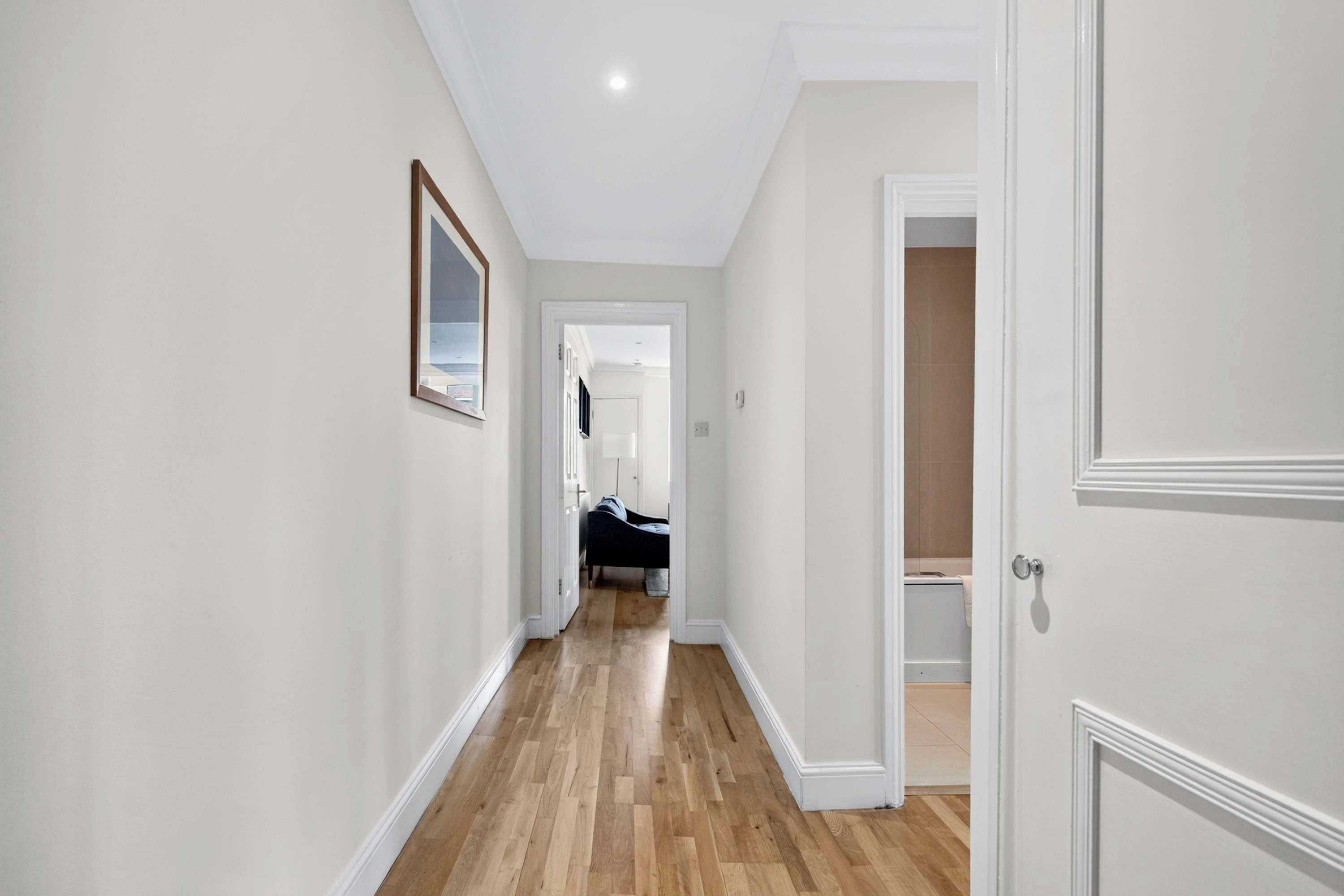 A hallway with wooden flooring, white walls, and a door leading to a room with a bed.