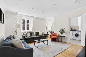 A modern living room with a sofa, armchair, coffee table, and a rug. There is a kitchen area with a washing machine and a dining table. - South Ken 2-bed - Science Museum - Lift - Sleeps 6 (London)