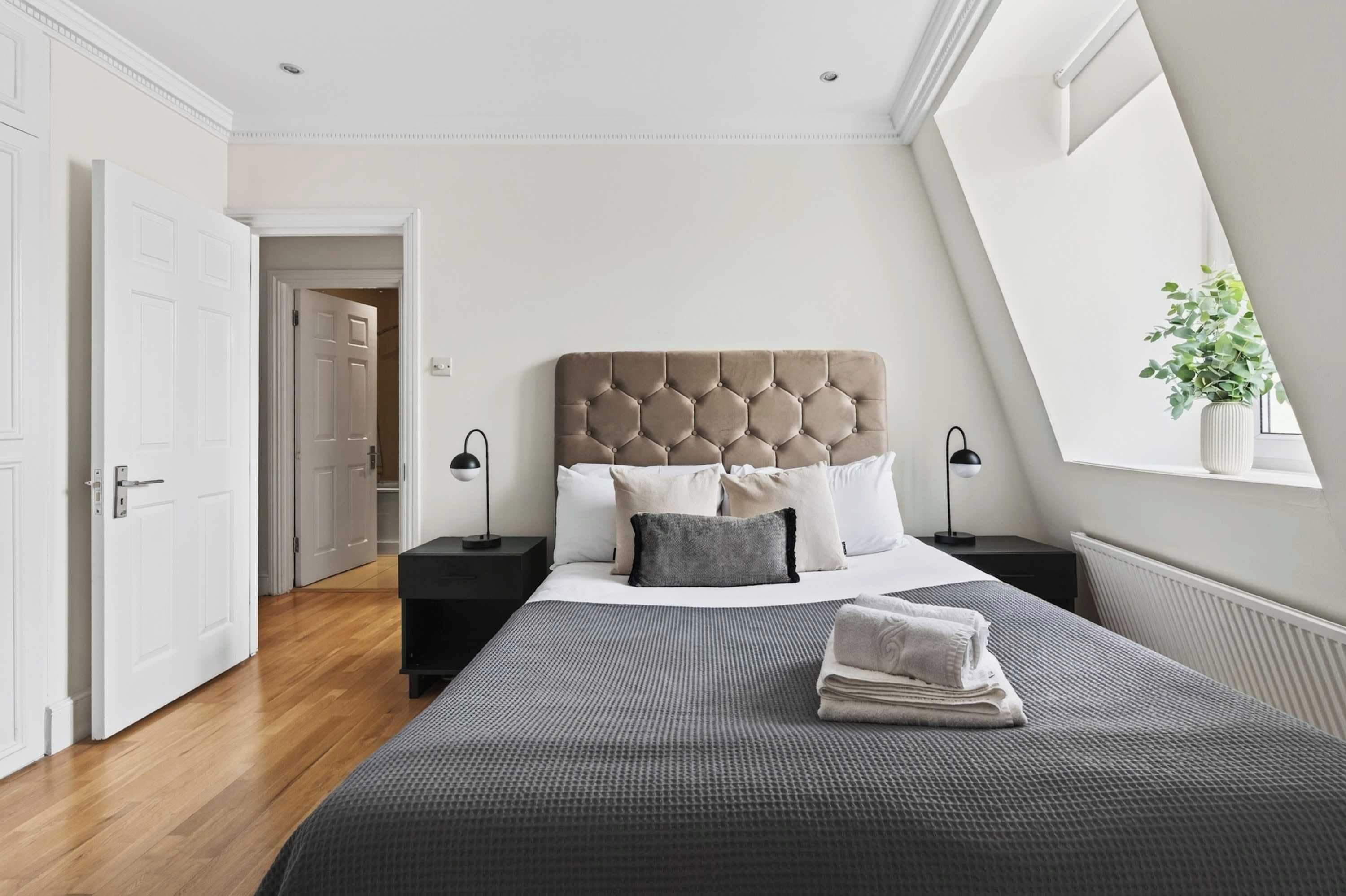 Un dormitorio moderno con una cama grande, dos mesitas de noche, una ventana con planta y una puerta que da a otra habitación.