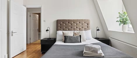 A modern bedroom with a large bed, two bedside tables, a window with a plant, and a door leading to another room.