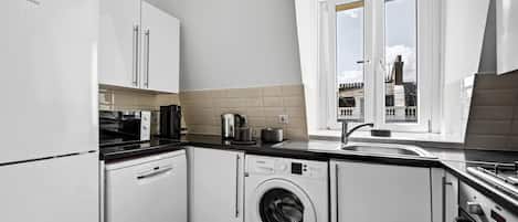 A compact kitchen with a built-in washing machine, a coffee maker, and a window with a view of the outside.