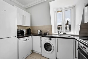 A compact kitchen with a built-in washing machine, a coffee maker, and a window with a view of the outside. - South Ken 2-bed - Science Museum - Lift - Sleeps 6 (London)