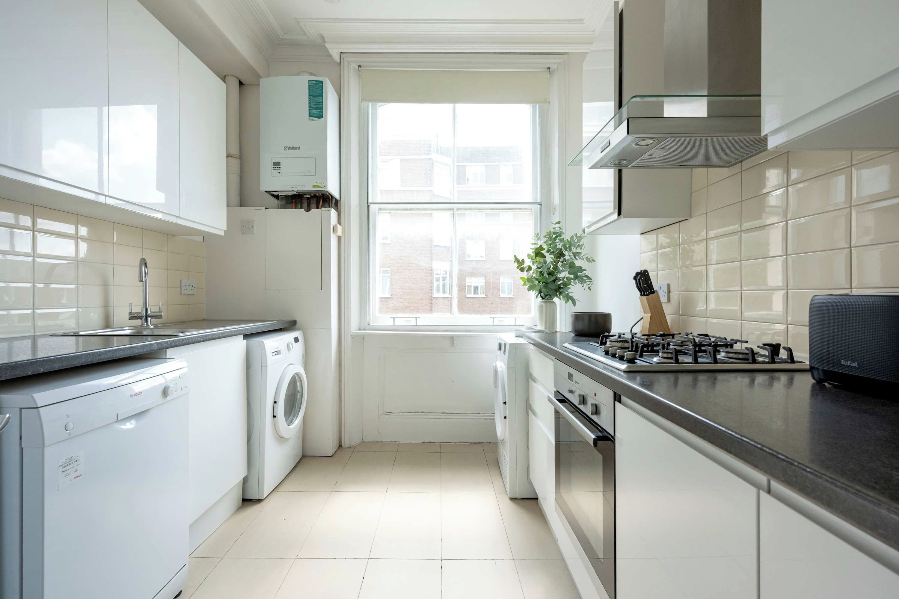 A modern kitchen with a dishwasher, washing machine, stove, and a window with a view of buildings.
