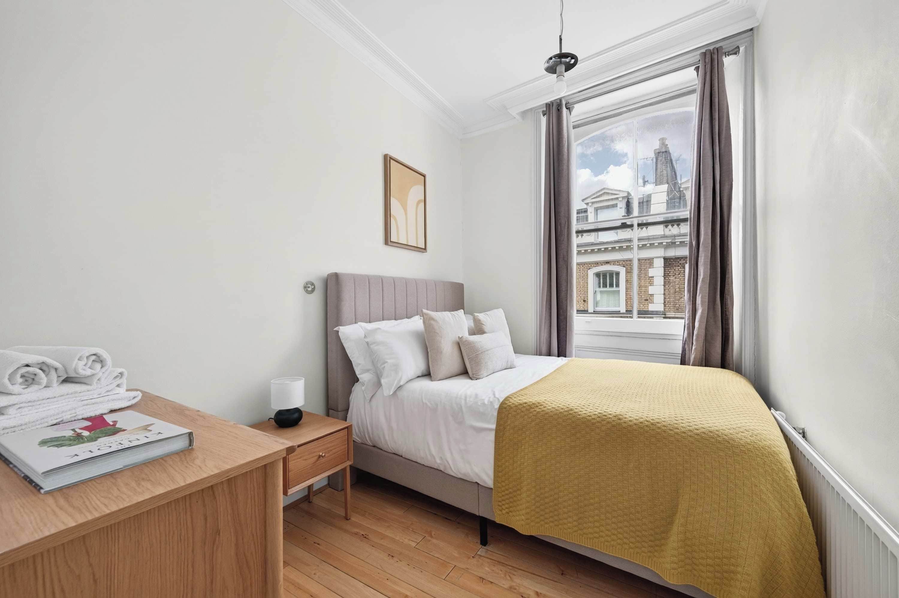 A bedroom with a bed, a bedside table with a lamp, a window with curtains, and a view of buildings outside.