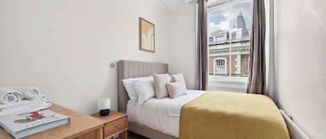 A bedroom with a bed, a bedside table with a lamp, a window with curtains, and a view of buildings outside.