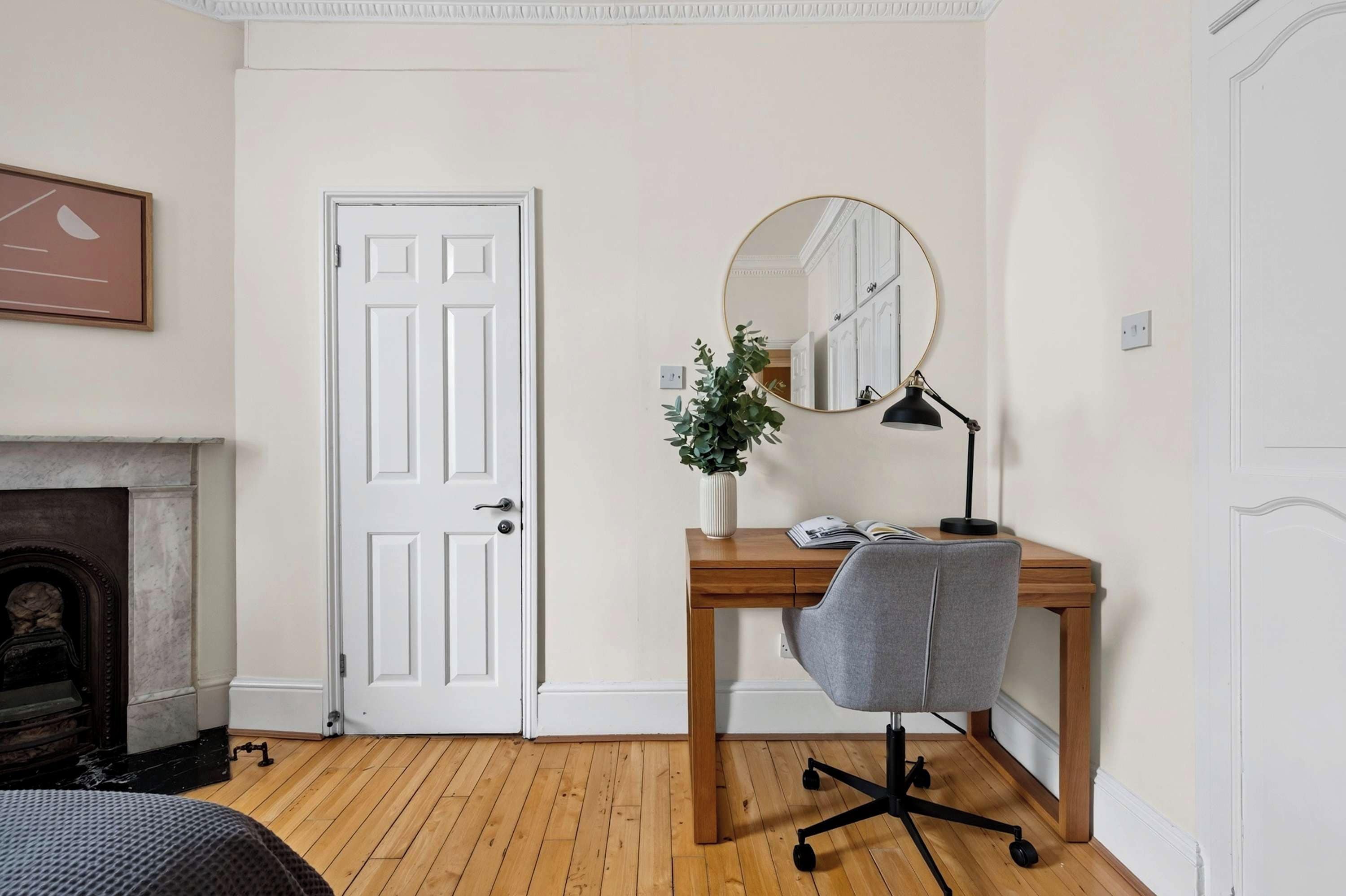 Una habitación con un escritorio de madera, una silla de oficina gris, un espejo redondo, una planta en maceta y una puerta blanca.
