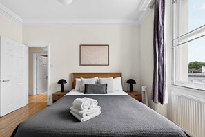 A hotel room with a large bed, a bedside table, a nightstand, a framed picture on the wall, and a window with curtains. - 2-bed by Natural History Museum - Lift - Sleeps 6 (London)