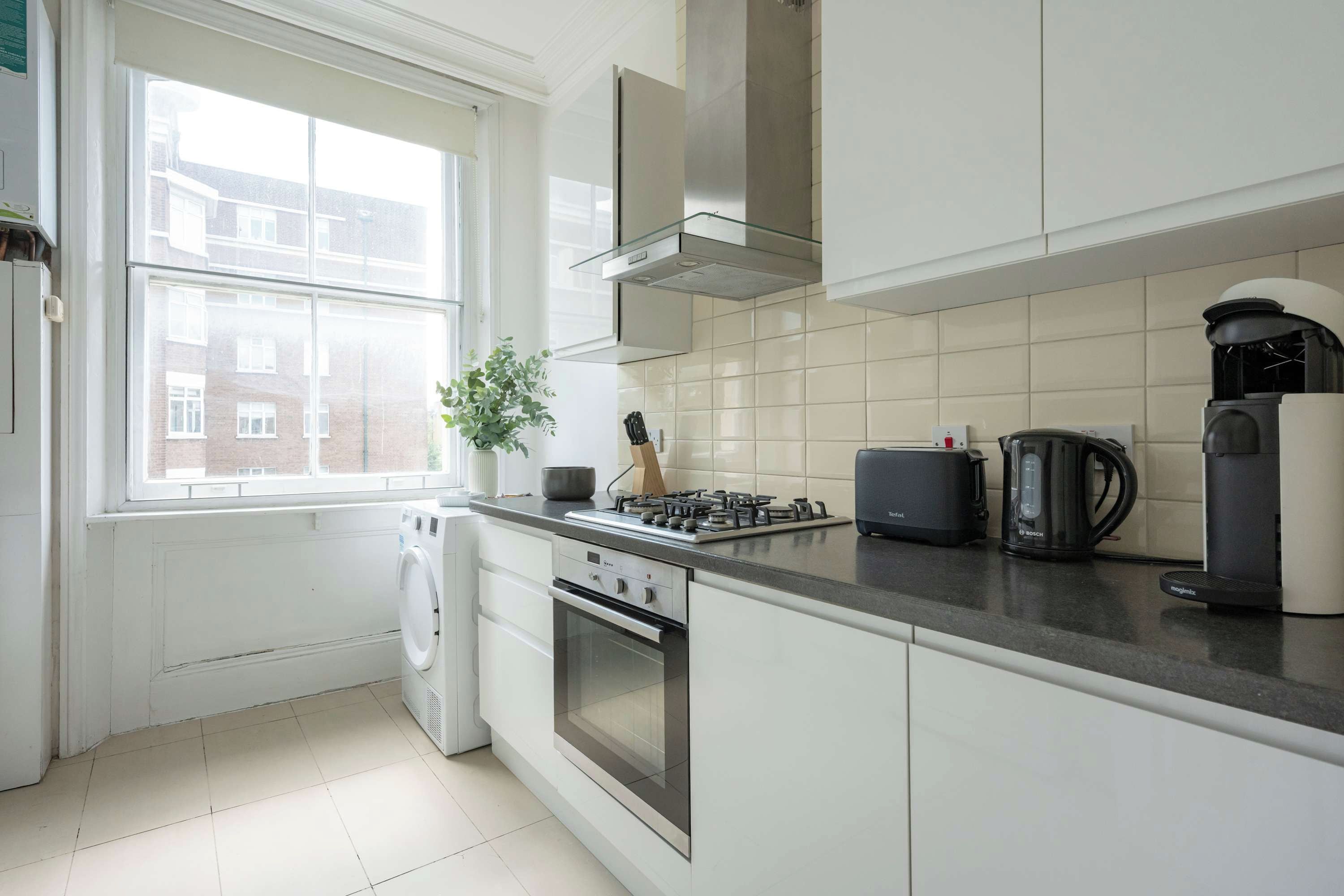 Une cuisine moderne avec des meubles blancs, une cuisinière à gaz, une machine à café Bosch et une vue sur un quartier résidentiel par la fenêtre.