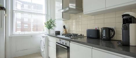Une cuisine moderne avec des meubles blancs, une cuisinière à gaz, une machine à café Bosch et une vue sur un quartier résidentiel par la fenêtre.