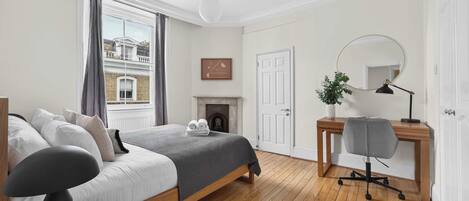 Un dormitorio con cama, escritorio, sillón, chimenea, ventana con cortinas y puerta.