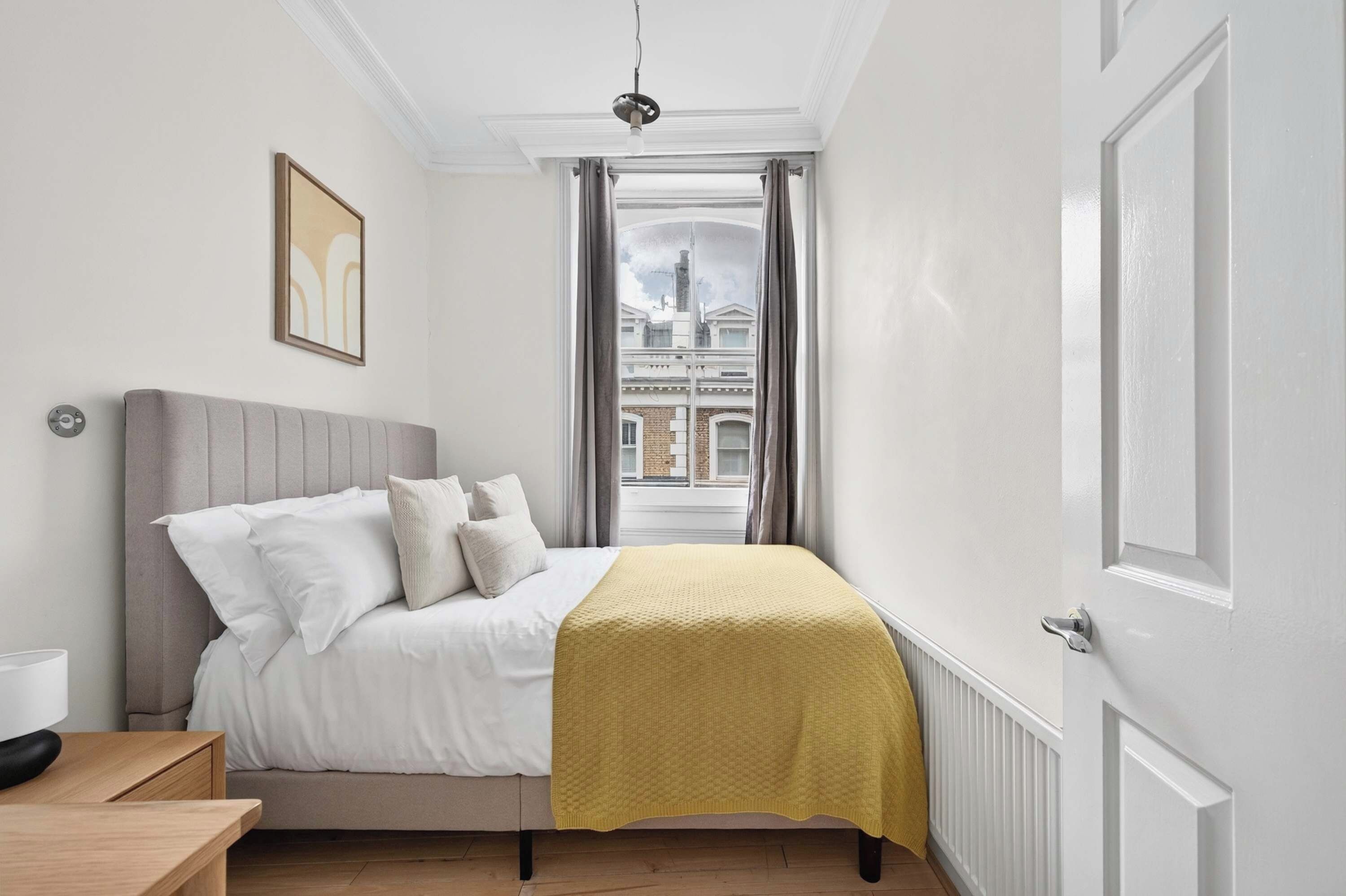 Ein Schlafzimmer mit einem Bett, einem Nachttisch, einem Fenster mit Vorhängen und Blick auf Gebäude.