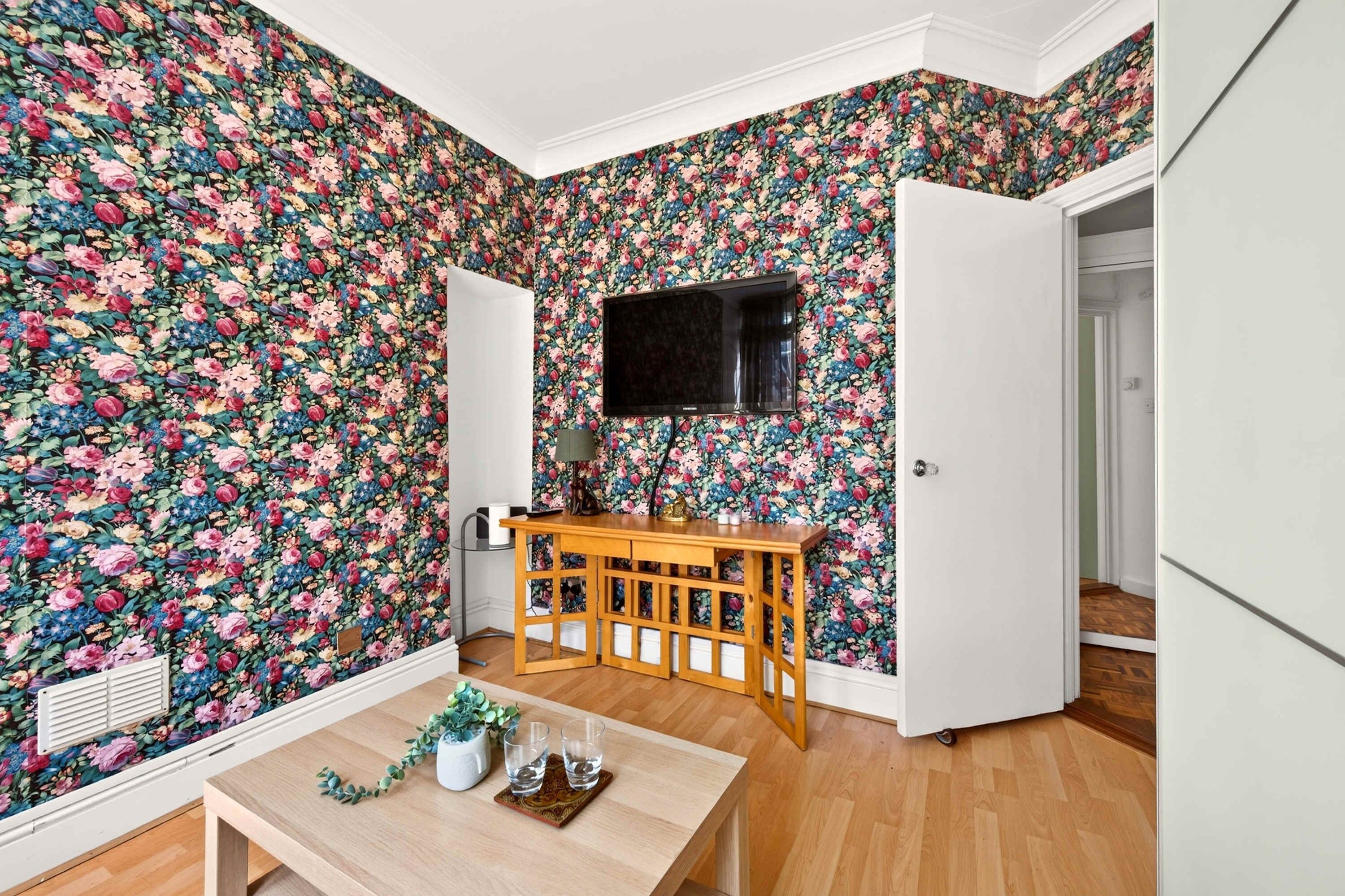 A room with floral wallpaper, a TV mounted on the wall, a wooden table with glasses and a plant, and a door leading to another room.