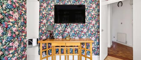 A room with floral wallpaper, a wooden table, a TV, and a radiator.