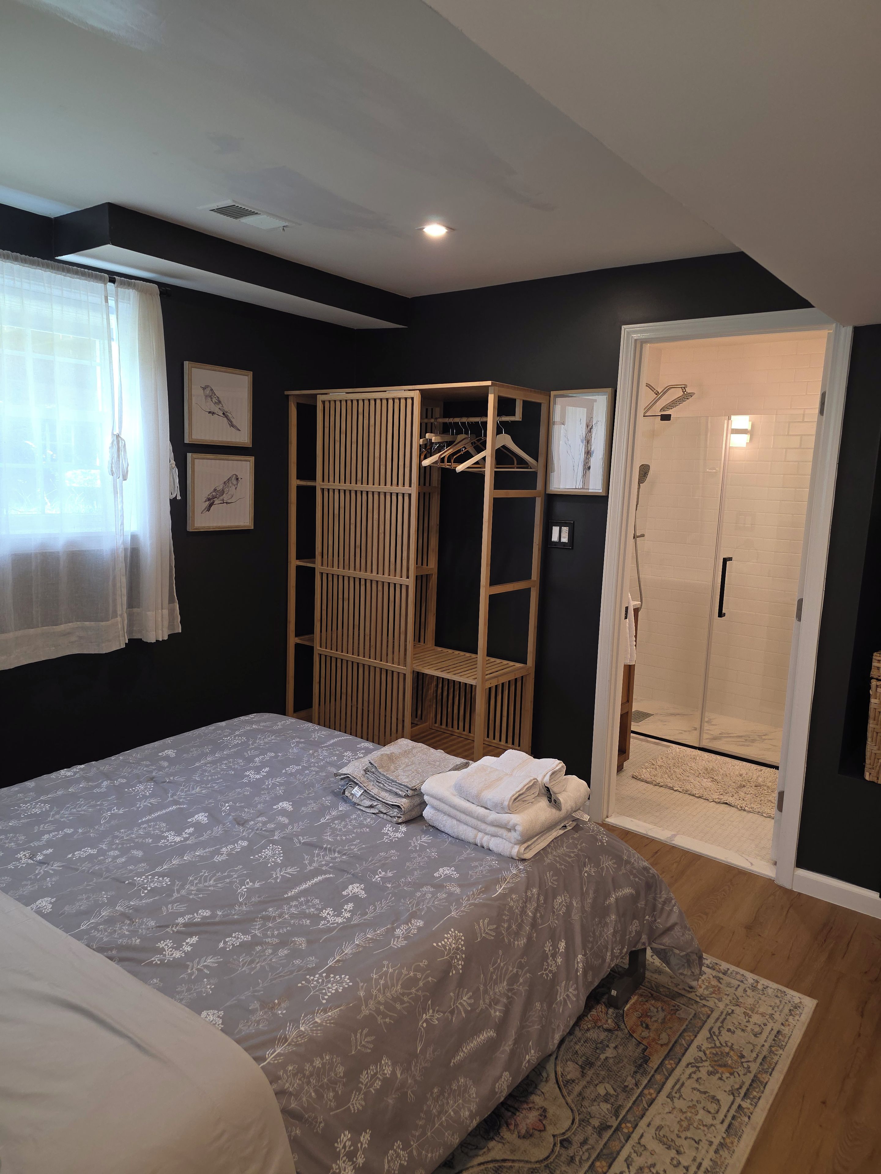 A bedroom with a bed, a wooden wardrobe, a shower, and a window with curtains.