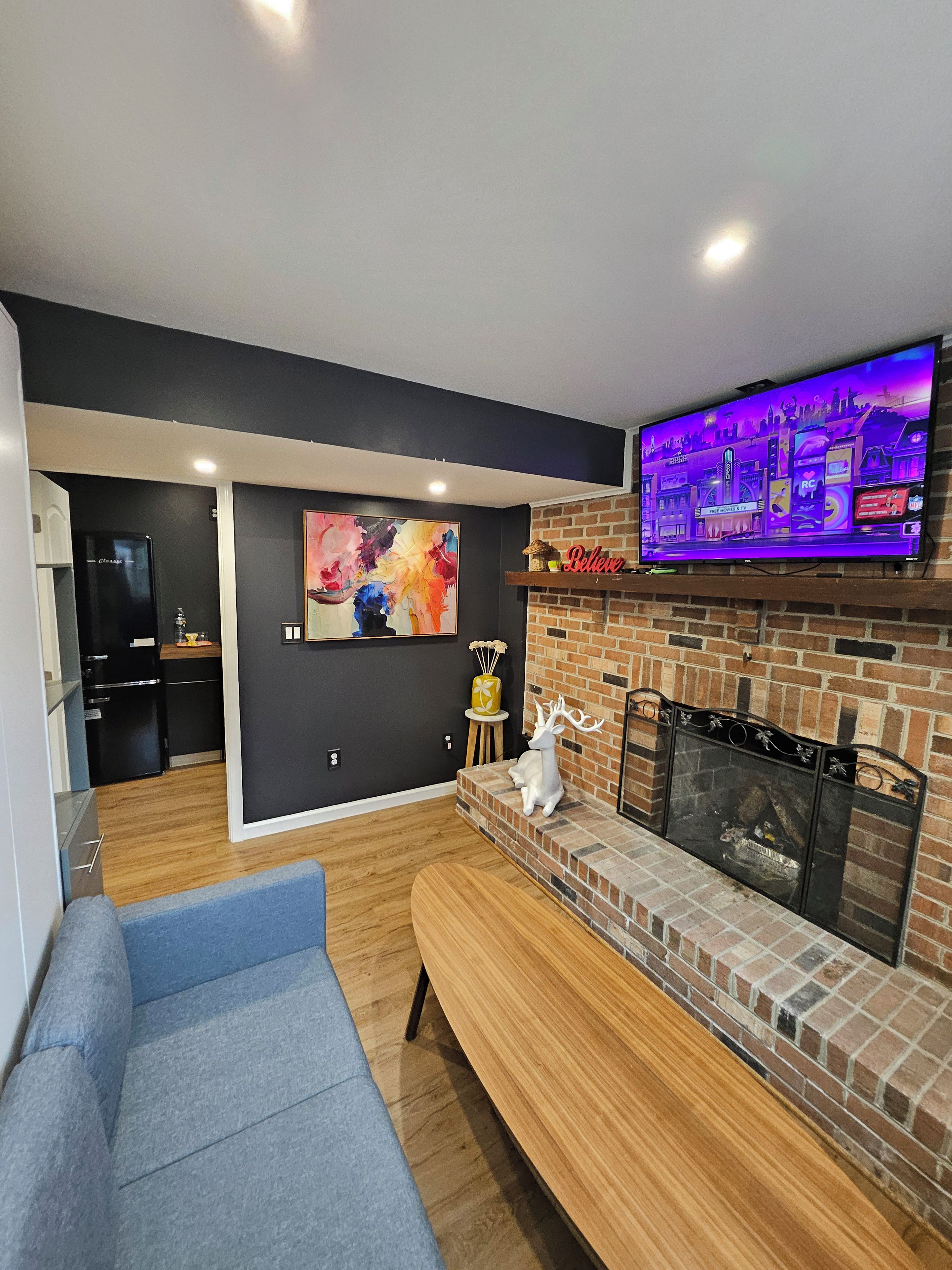 A modern living room with a flat-screen TV, a fireplace, a blue sofa, and a kitchen area in the background.