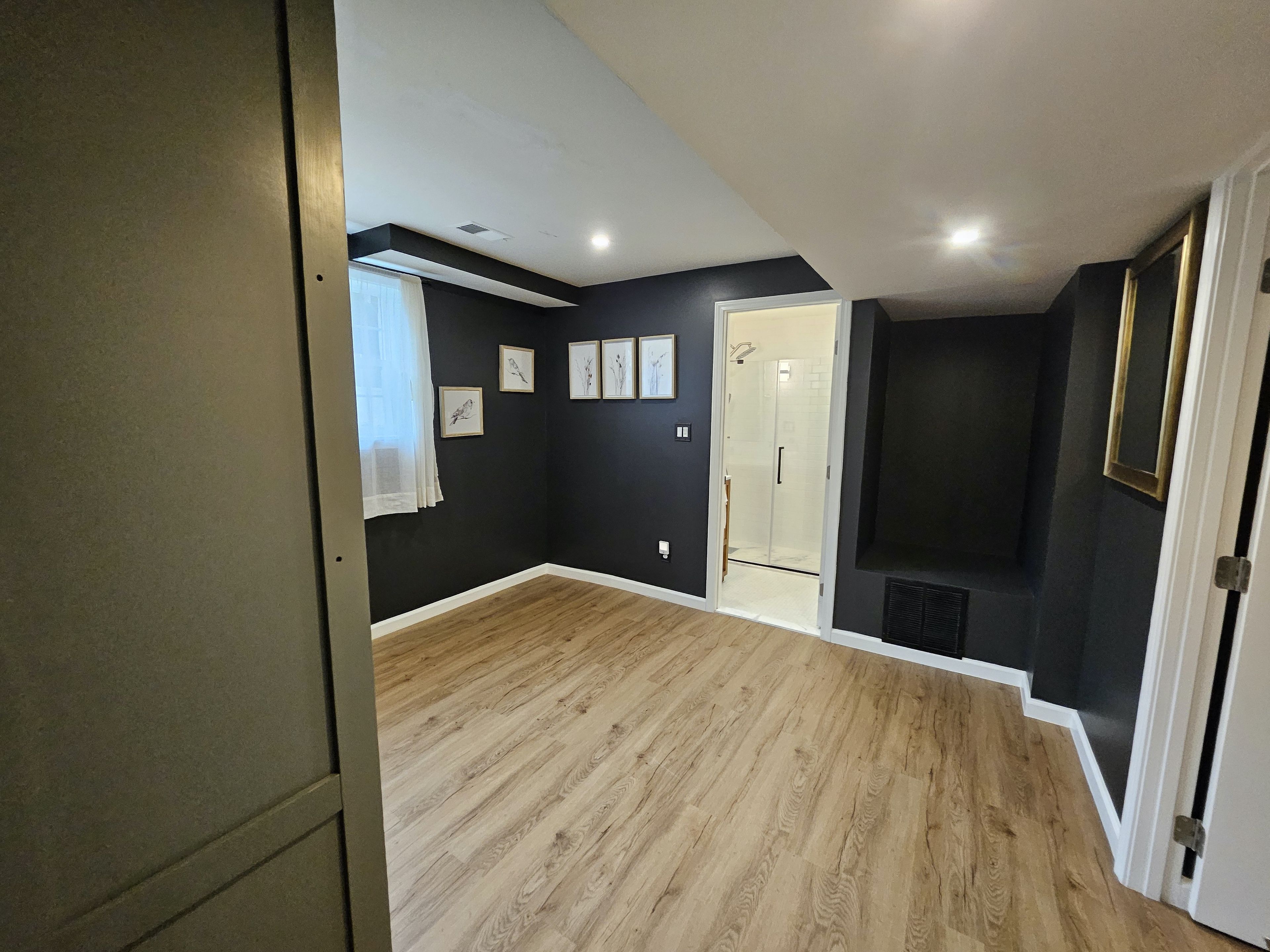 A room with a wooden floor, a door, a window with curtains, and a wall with framed pictures.