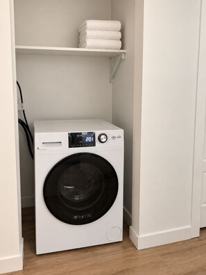 A compact, white front-loading washing machine in a small laundry room with a shelf above it holding folded towels. - Midtown Gem Near Downtown (Detroit)