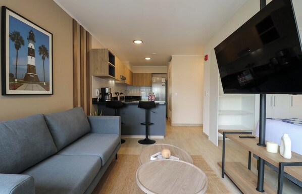 A modern living room with a grey sofa, a coffee table, a TV, and a kitchen in the background. - w* | Charming Loft w/ Balcony in Prime Miraflores (Miraflores)