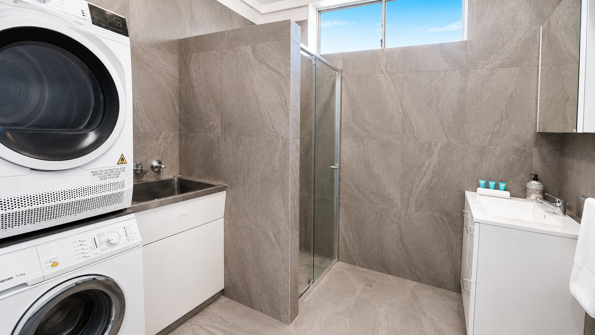 A modern laundry room with a front-loading washer, a sink, and a glass-enclosed shower.