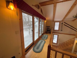 Un intérieur en bois, de grandes fenêtres, un escalier et une vue sur une terrasse enneigée à l’extérieur.