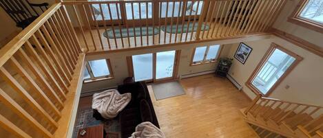A spacious loft with a high ceiling, a wooden staircase, a cozy seating area with a sofa and chairs, and large windows offering a view of a snowy landscape.
