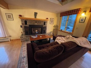 Un salon chaleureux avec une cheminée en pierre, un canapé marron, une table basse et une grande fenêtre donnant sur l’extérieur.