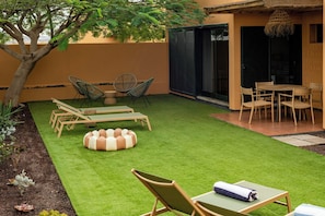 Une terrasse avec une pelouse artificielle verte, des fauteuils de jardin en bois, un pouf rond et une table à manger avec des chaises.