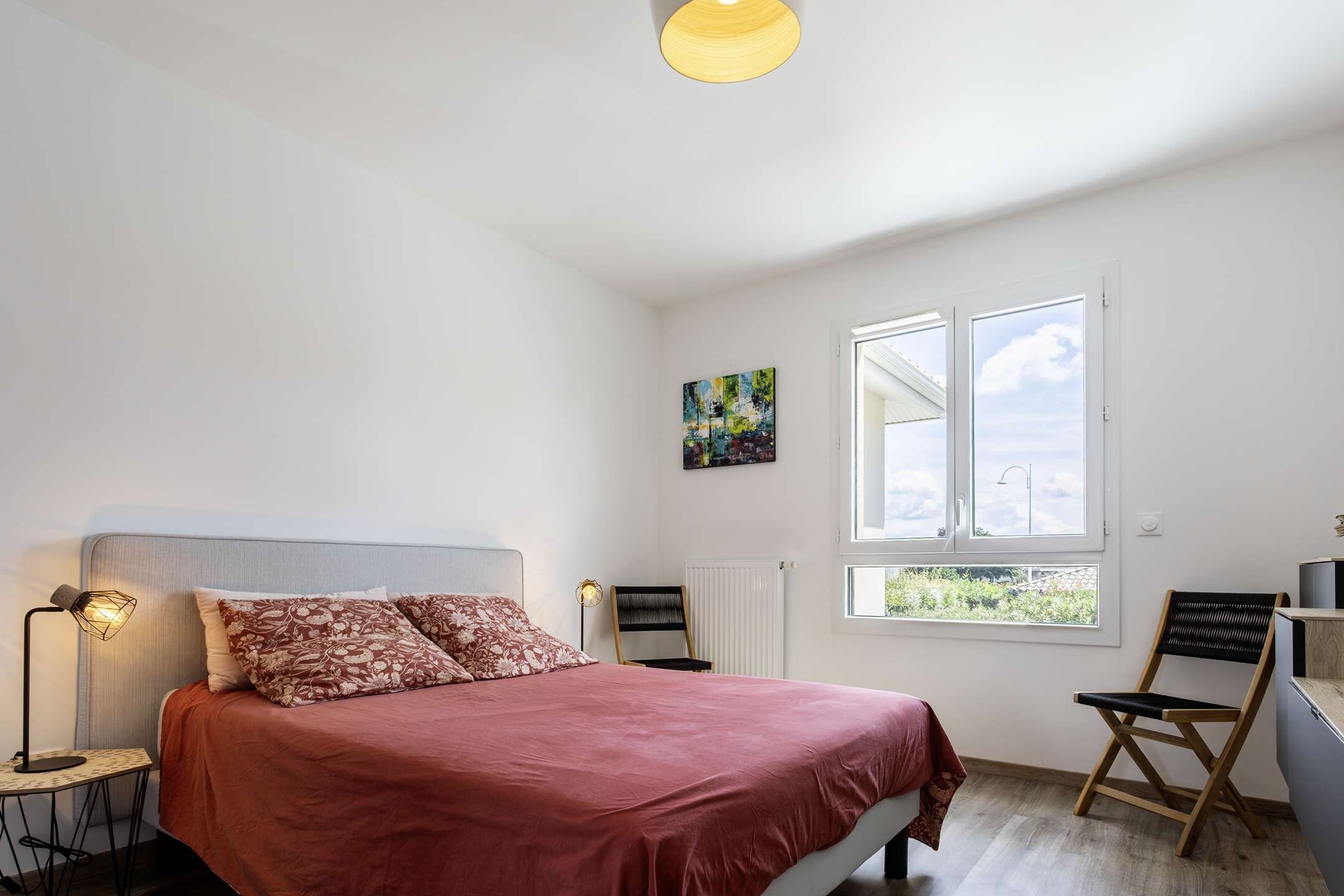 Un dormitorio con cama, una silla, una mesa, una lámpara, un radiador y una ventana con vistas.